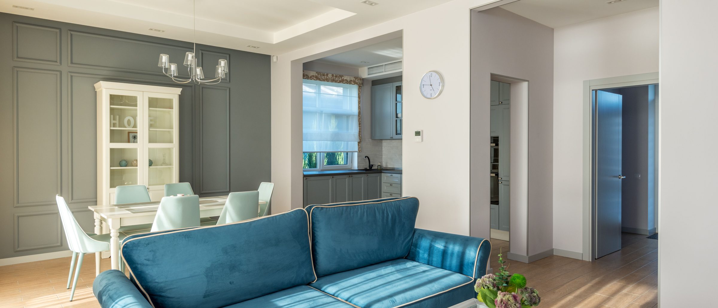 A furnished open-plan living and dining room with a teal velvet sofa, dining table and view through to the kitchen